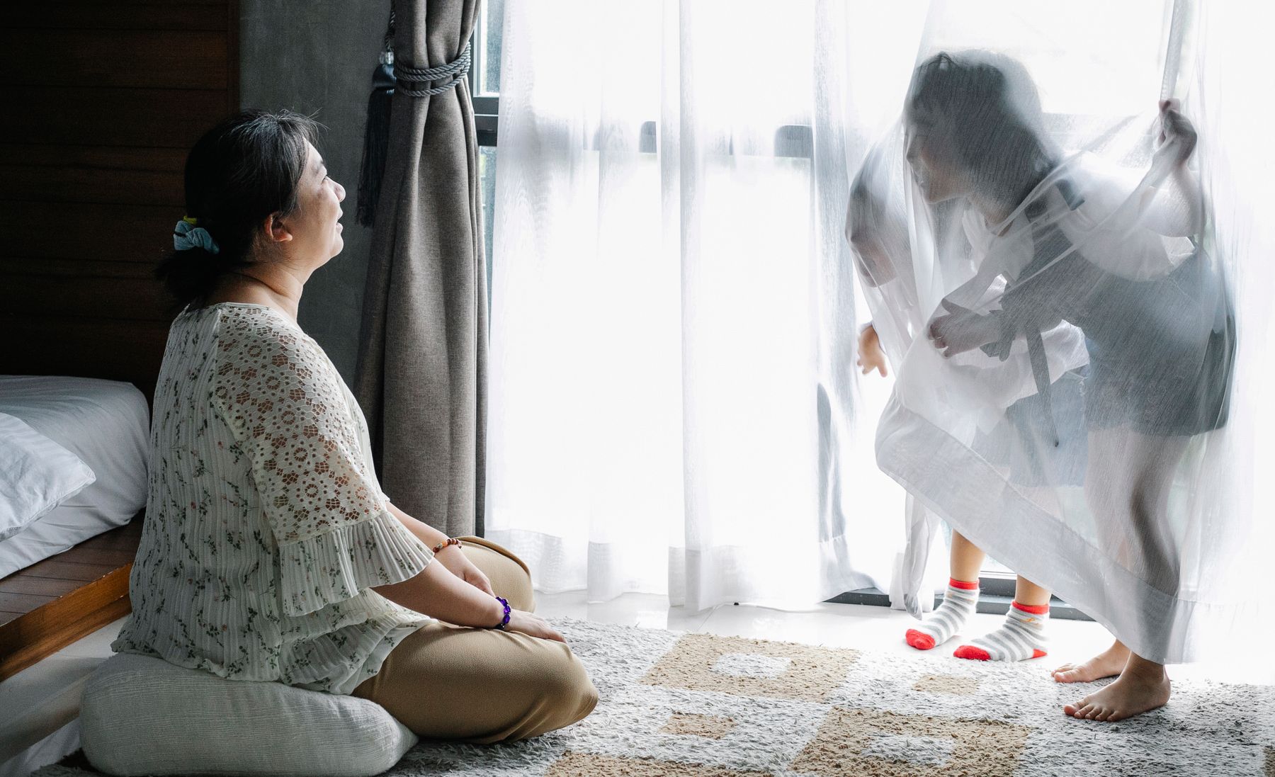 Seorang ibu berehat di rumah dalam cahaya pagi sementara anaknya bermain di tepi tingkap